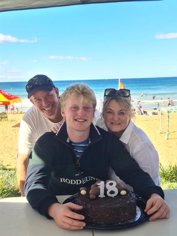 Three people at a beachside celebration with a chocolate cake marked "18".