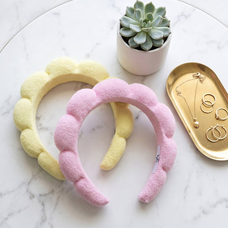 Two fuzzy headbands, one pink and one yellow, on a marble surface with a small plant and jewelry dish.